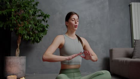 Zoom-in-Confident-brunette-girl-in-a-gray-top-and-green-sweatpants-does-yoga-sits-in-the-lotus-position-and-practices-breathing-practices-in-a-modern-apartment-at-home-near-a-houseplant