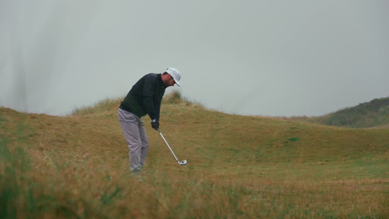 Male golfer hits golf ball wedge shot from tall grass rough in the rain on Ireland links golf course with water splashing in slow motion