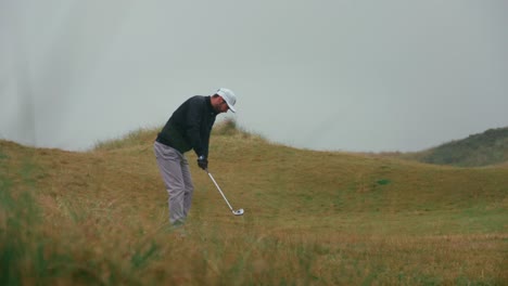 male golfer hits golf ball wedge shot from tall grass rough in the rain on ireland links golf course with water splashing in slow motion