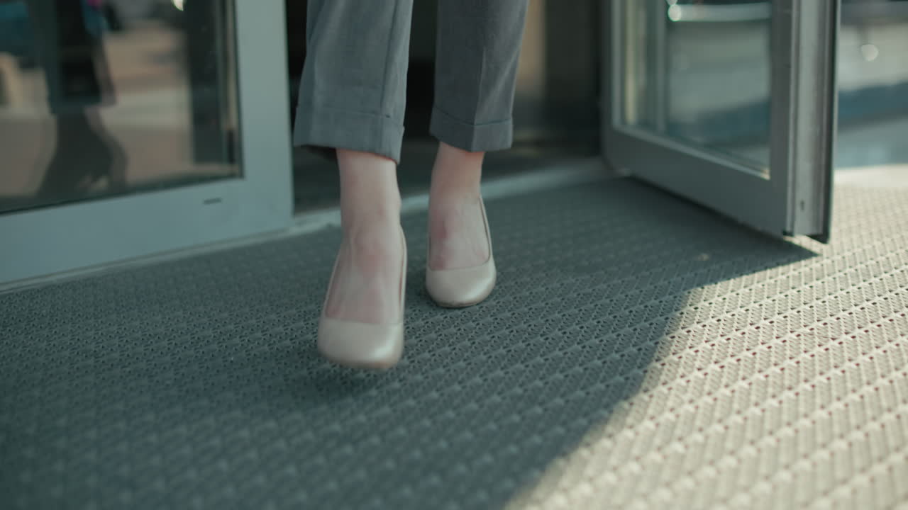 Lower angle view of professional woman in heels stepping out of building through glass door, focus on movement, footwear, modern office environment