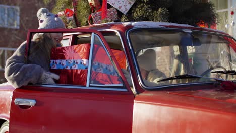 happy little girl putting new year present box into car with decorated holiday tree. cheerful girl delivering christmas gifts in car on holiday eve. happy winter new year mood and christmas spirit.