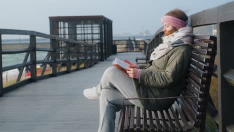 woman reading a book on a wooden bench at the beach