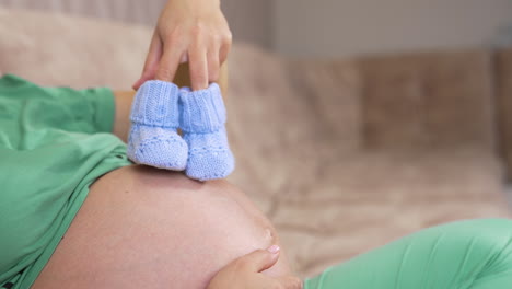 pregnant woman holding baby booties at home, closeup. eight months pregnant woman with blue booties on her belly
