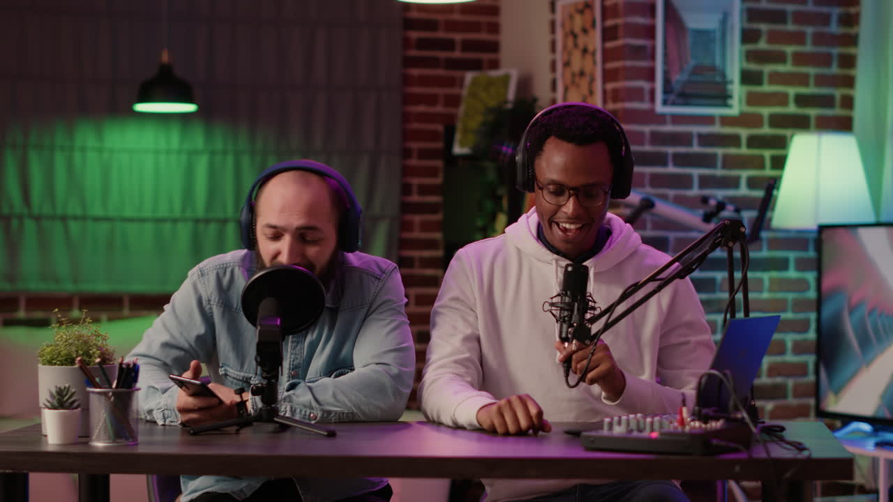 Diverse men in dual vlog recording review of digital smartphone in podcast studio with professional microphone