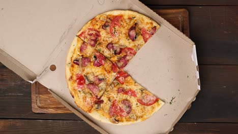 baked round pizza with cheese, tomatoes, sausage and mushrooms cut into pieces in a white cardboard box, female hand takes a slice