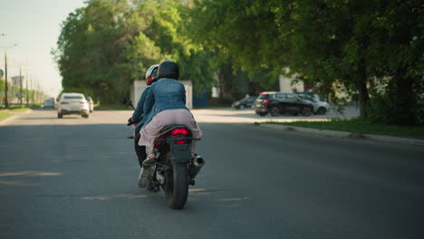 two women ride a power bike on an urban road. the passenger holds onto the rider as they pass a pedestrian signpost, trees line the road, with an electric pole and a blurred car ahead of them