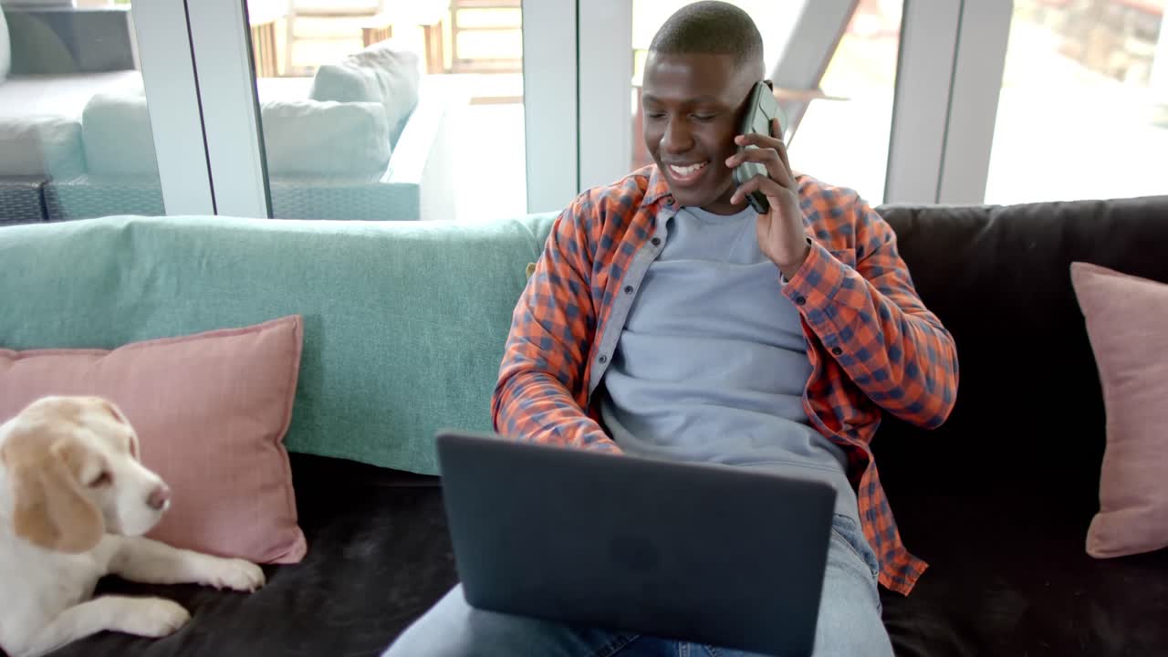 Happy african american man on smartphone and using laptop at home, with his pet dog, slow motion