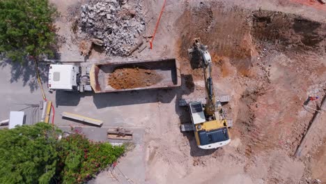 Von-Oben-Nach-Unten-Luftaufnahme-Eines-Bulldozers,-Der-Sand-Schaufelt-Und-Ihn-Auf-Einer-Baustelle-In-Den-LKW-Füllt