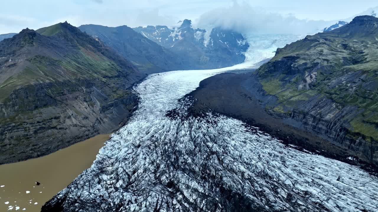 Dronebillede i fugleperspektiv af en islandsk gletschertunge, der flyder gennem en vulkansk dal mod en mudret smeltevandslagune med grønne bjerge og mørke morænekanter