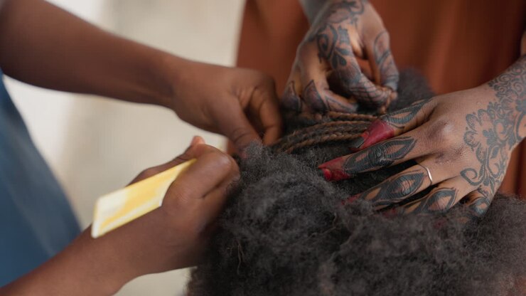 Intricata acconciatura a treccia su cliente nero con mani tatuate, primo piano di afro texturizzato e unghie lunghe dipinte, dita che intrecciano piccole sezioni con ritmo preciso, una scena intima di cura artistica.