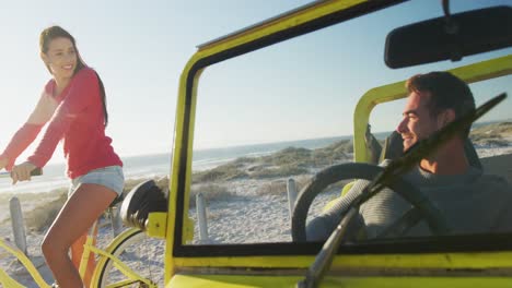 feliz mujer caucásica montando una bicicleta al lado de un hombre en un buggy de playa junto al mar