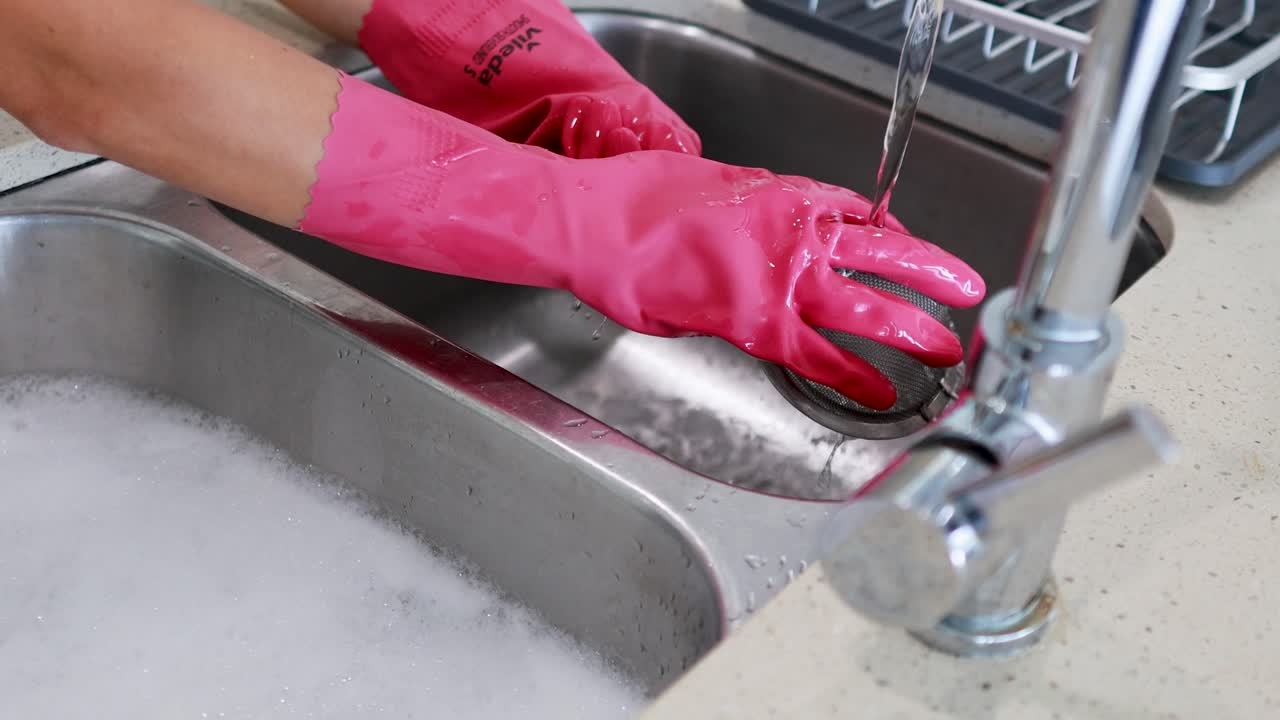 Hands in pink gloves wash dishes in a bright kitchen sink under running water