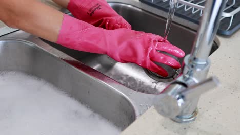 hands in pink gloves wash dishes in a bright kitchen sink under running water