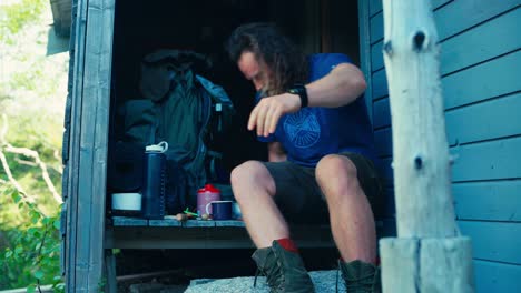 man sits on doorway of wooden cottage with breakfast and coffee prepared in morning