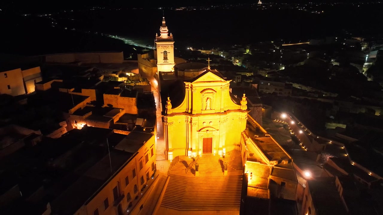 Una chiesa luminosa si erge sopra il centro storico di notte, vista da un drone sulla città costiera