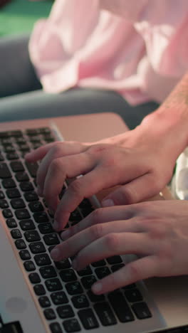 primer plano de manos escribiendo en el teclado de una computadora portátil al aire libre mientras otra persona sostiene un libro en el fondo, con vegetación borrosa y luz solar