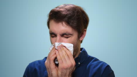 Sick-man-sneezing-in-studio.-Ill-man-using-napkin-on-blue-background