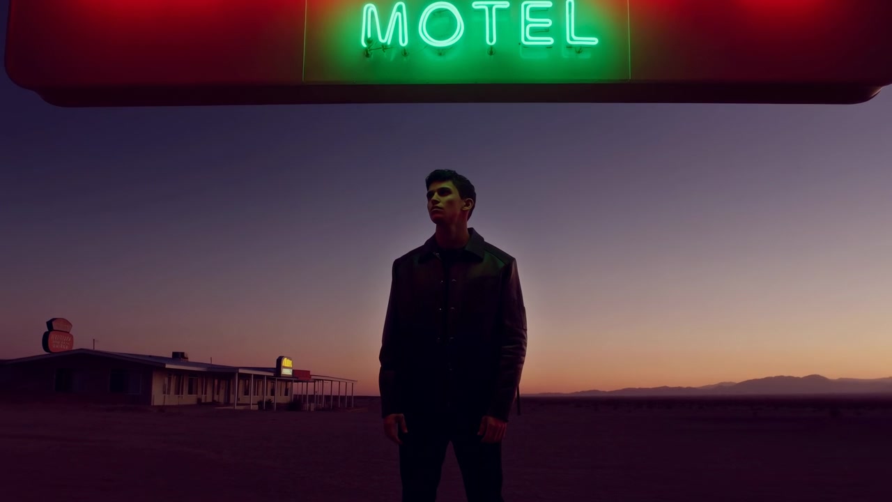 Homme sous une enseigne de motel néon au crépuscule, un vagabond solitaire se tient au premier plan, veste en cuir rétroéclairée par des lumières rouges et vertes.