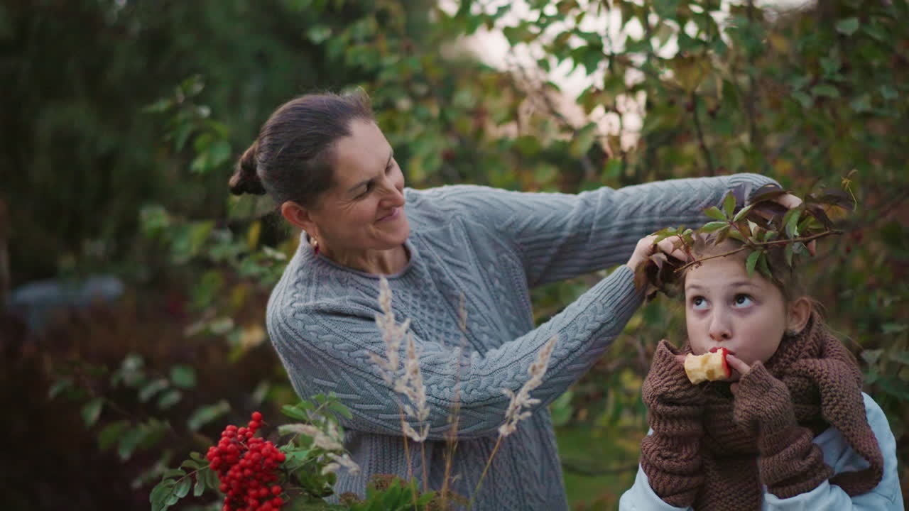 Eine Frau bastelt mit einem Kind im Garten bei Dämmerung eine Blattkrone. Sie legt Laub und Beeren auf den Kopf des Kindes, während dieses einen kleinen Snack hält. Gemütliche Strickpullover, spielerische Verbundenheit und eine warme, saisonale, authentische Atmosphäre.