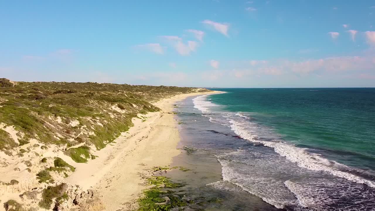 An empty coastline with waves crashing along a sandy beach under clear skies, aerial view