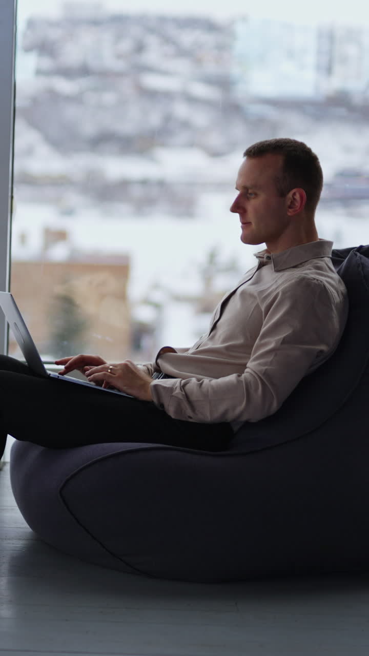 Freelance male businessman working at laptop in comfortable room. Man sitting in bean bag chair at backdrop of panoramic windows and winter cityscape in blur. Vertical video