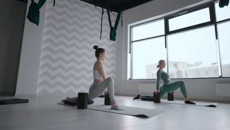 Grupo-De-Mujeres-Jóvenes-Estirándose-En-Un-Gimnasio-Con-Ventanas.-Dos-Atractivas-Chicas-Deportivas-Practican-La-Pose-De-Yoga-En-Barco-Nauka-Asana-Sobre-Una-Alfombra-Negra-En-Clase-De-Fitness.-Dos-Mujeres-Estirando-Las-Piernas-Juntas-En-El-Estudio-De-Yoga