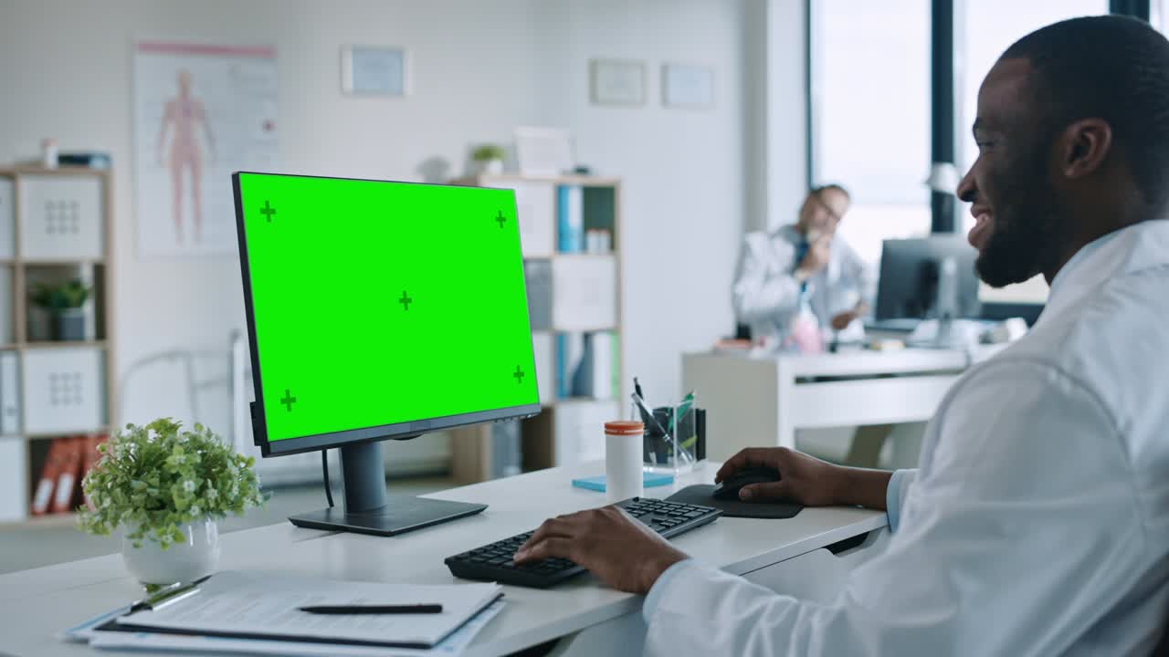 un medico afroamericano sta lavorando su un computer con schermo verde in una clinica sanitaria. un assistente in camicetta bianca sta leggendo la storia medica dietro una scrivania nell'ufficio dell'ospedale.