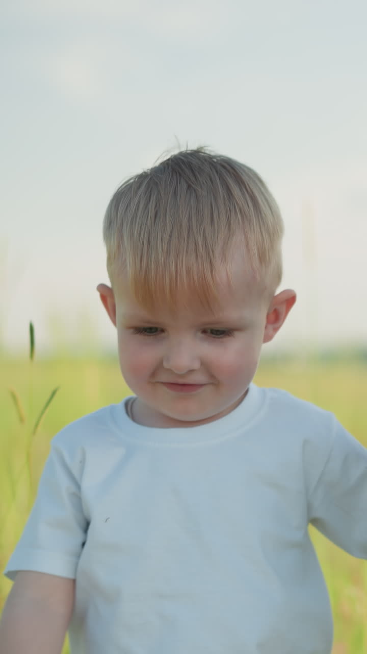 крупный план молодого мальчика в белой рубашке, улыбающегося, когда он идет по травяному полю, держа руку своей матери. мать, одетая в синее платье, имеет ее лицо вне поля зрения