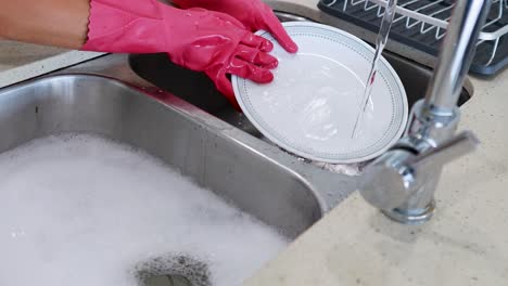 hands in pink gloves wash dishes under running water in a bright kitchen setting