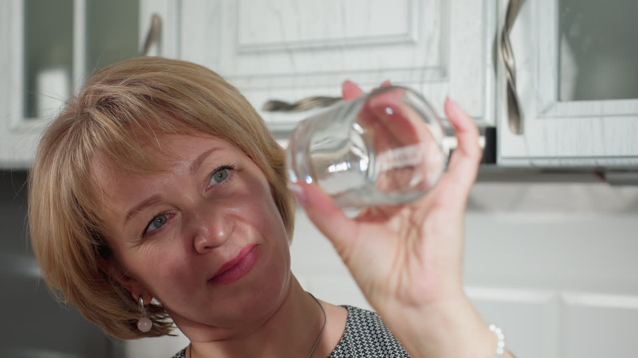 Lady carefully observing sparkling clean transparent glass cup held against light in bright modern kitchen with white cabinets, ensuring it is spotless and polished to perfection after cleaning