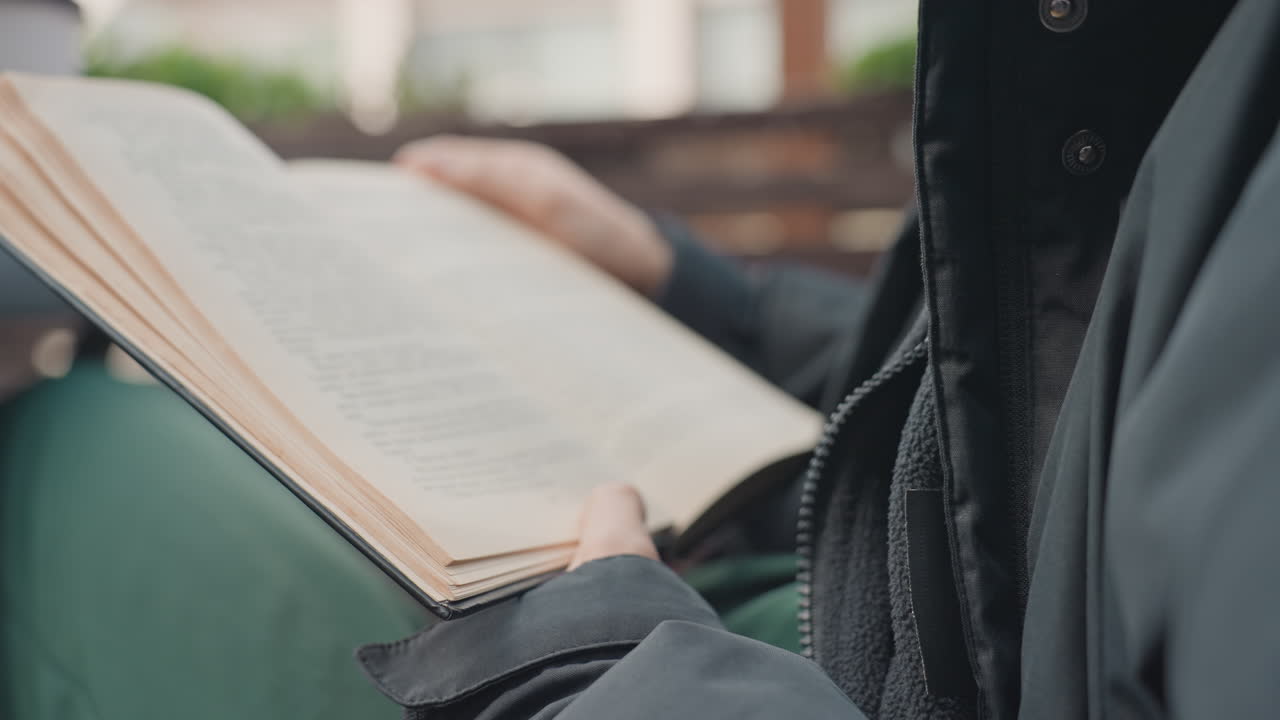 mani che sfogliano le pagine di un libro su una panchina, luce naturale soffusa, libro tascabile usato, giacca a strati, listelli di legno, atmosfera lenta e riflessiva, dita che tracciano le righe, ambientazione autunnale esterna, profondità di campo ridotta