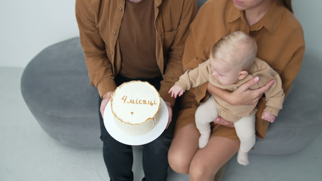 Man holds a big white cake with a sign "four months". Little baby in woman's hands tries to touch the dessert. Top view.