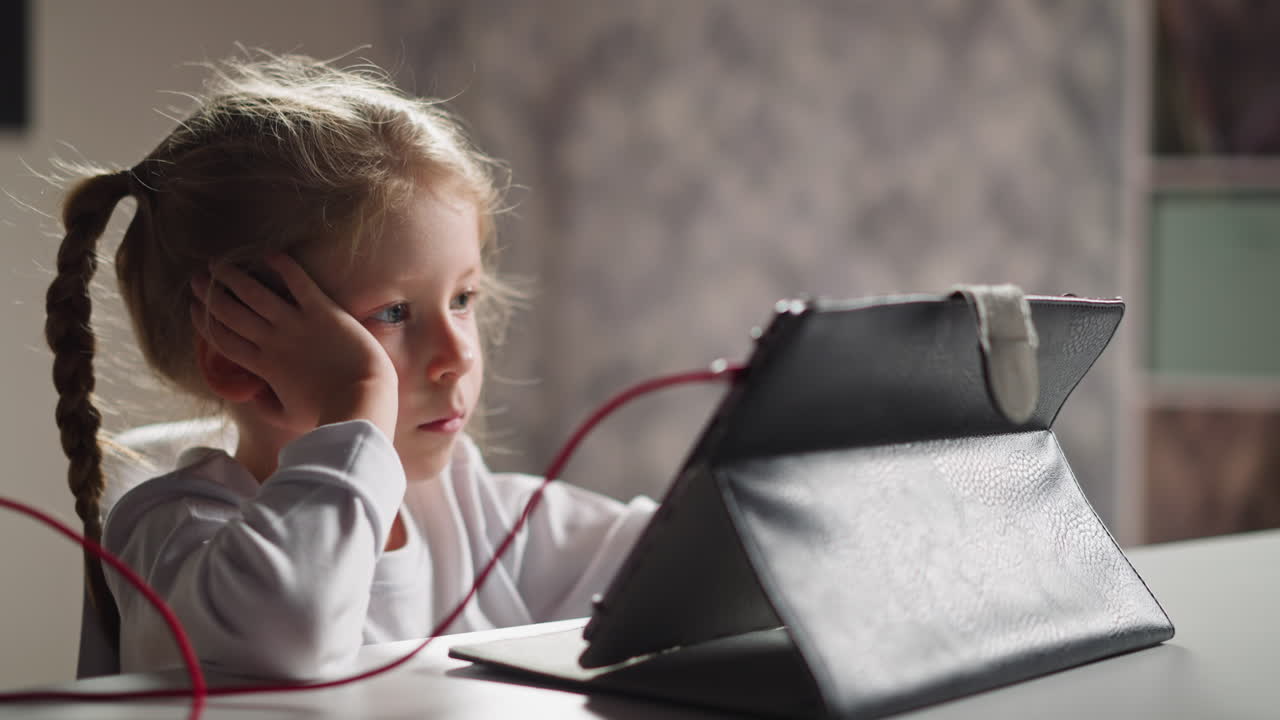 Una bambina annoiata guarda un video educativo tramite tablet al tavolo.