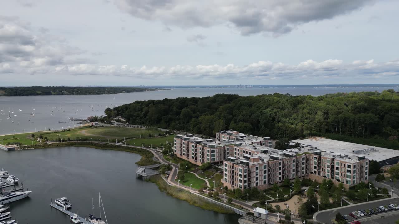 Vista aérea panorâmica afastando-se de uma grande marina repleta de iates e veleiros, revelando uma bela costa verdejante, uma baía e um horizonte urbano distante sob um céu nublado.