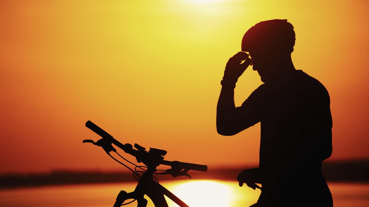 Professional cyclist takes off his protective glasses while standing at a beautiful sunset. Silhouette of a sporty cyclist in helmet on a bike taking down glasses at golden color of sun setting.