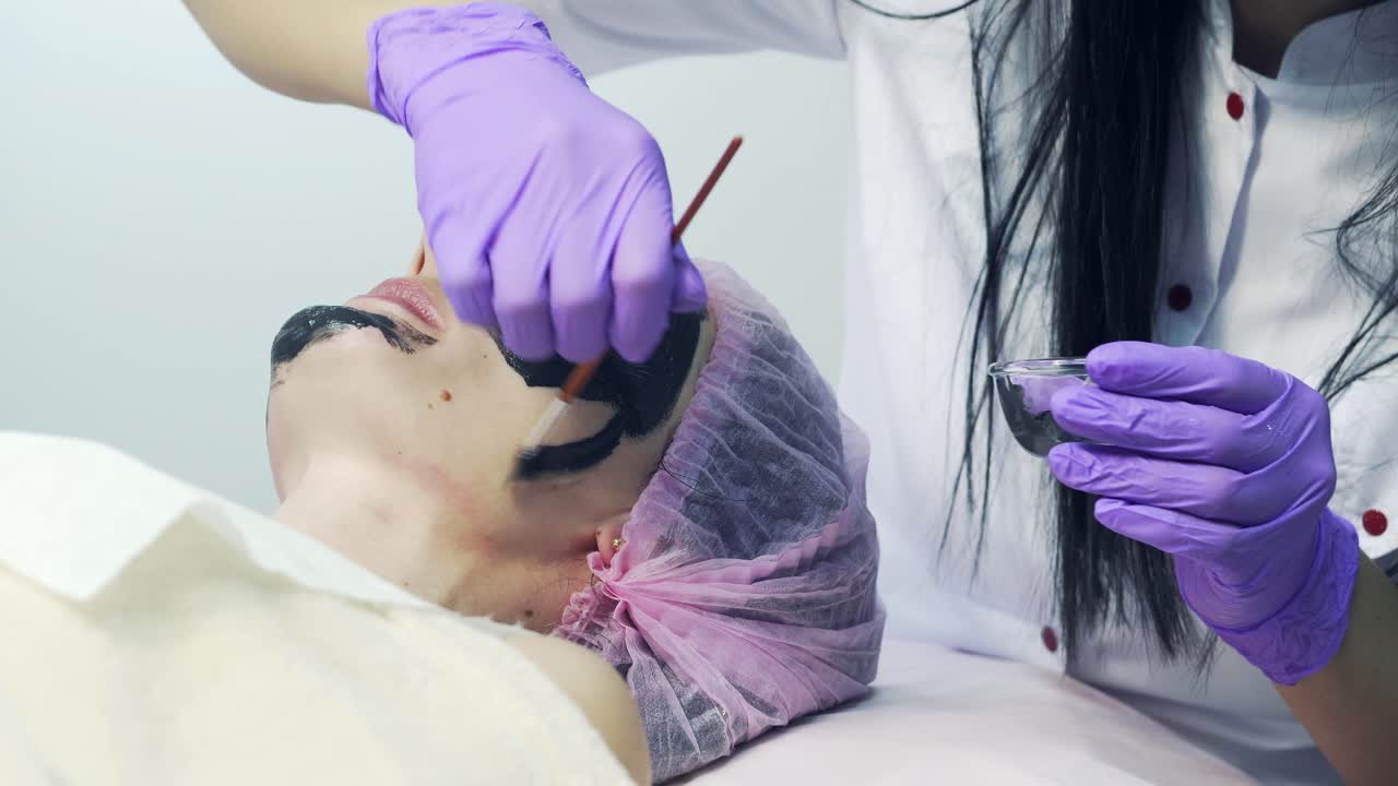 Beautiful young woman is having facial mask while lying down in spa. Female is getting mud black mask cosmetic procedure by cosmetologist.