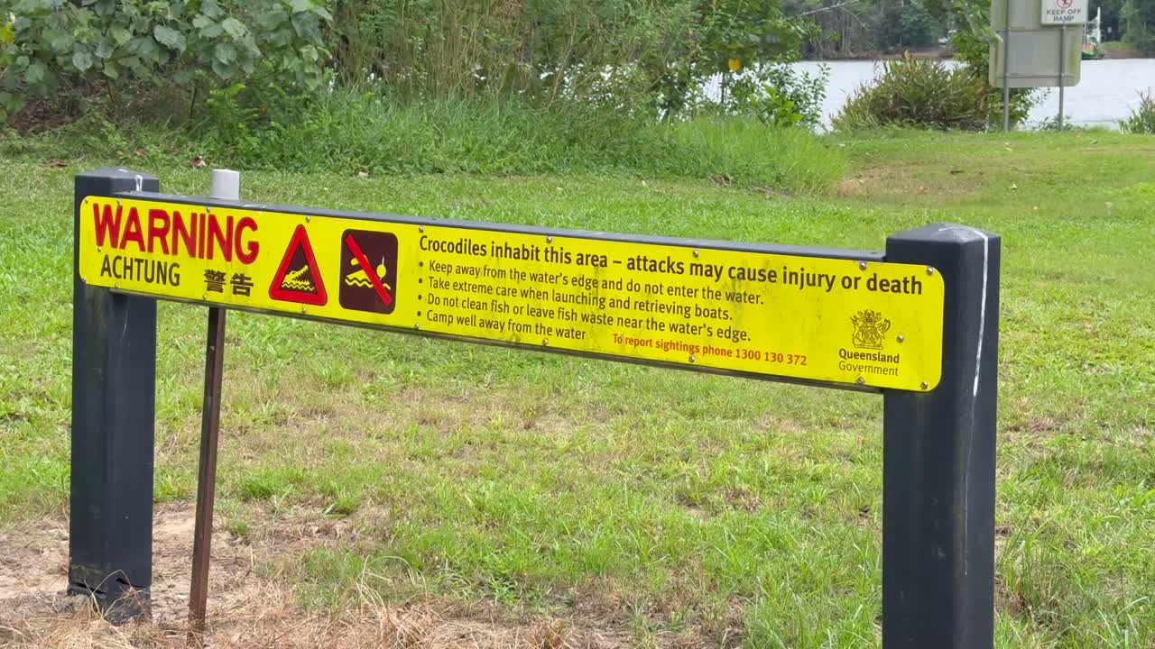 A stationary camera captures a bright yellow warning sign about saltwater crocodile danger by a grassy riverbank in daylight, with minimal movement