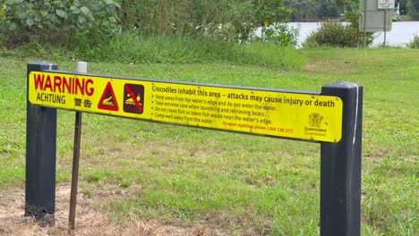 a stationary camera captures a bright yellow warning sign about saltwater crocodile danger by a grassy riverbank in daylight, with minimal movement