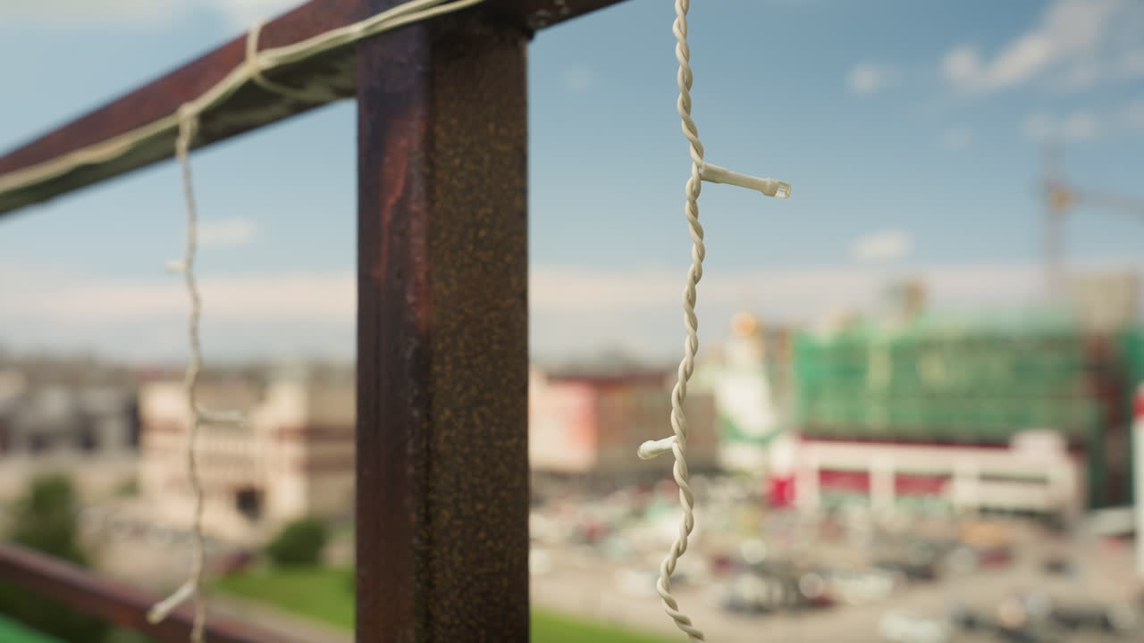 Đèn treo lan can ban công nhìn ra cảnh quan thành phố, hiệu ứng bokeh điện ảnh và độ sâu trường ảnh nông, cột kim loại rỉ sét với lớp sơn bong tróc, dây điện lủng lẳng đung đưa trong gió nhẹ, các tòa nhà và bãi đậu xe bị mờ