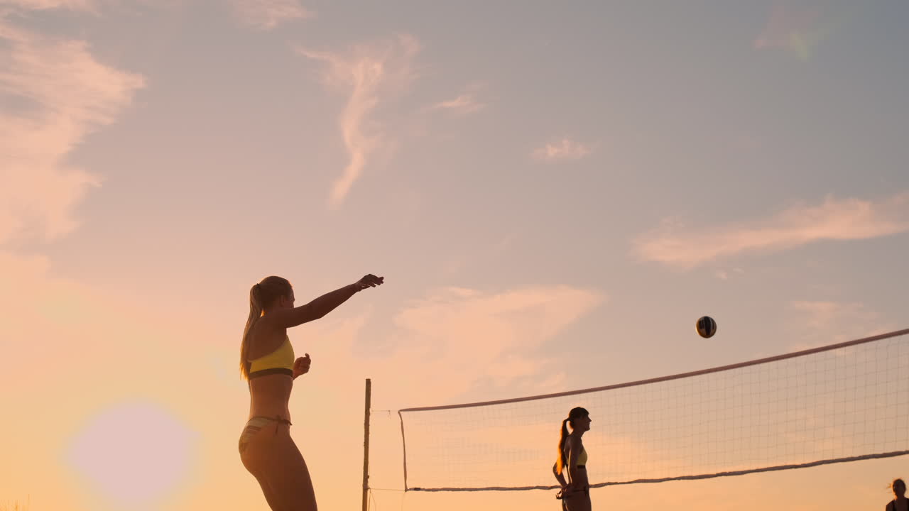 해변 볼리볼을 하는 운동선수 소녀는 공중으로 뛰어내리고 아름다운 여름 저녁에 네트 위에 공을 때립니다.