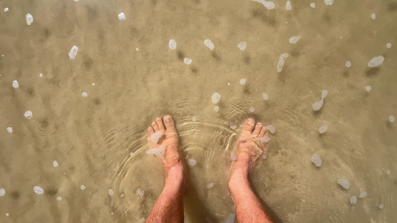 젊은 남자가 바닷물에 발을 담근 채 해변 모래사장에 서 있어요