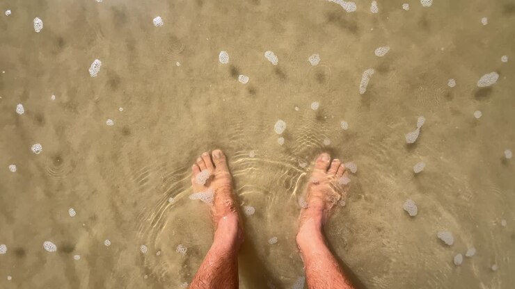 Giovane uomo in piedi sulla sabbia della spiaggia con l'acqua del mare che gli bagna i piedi