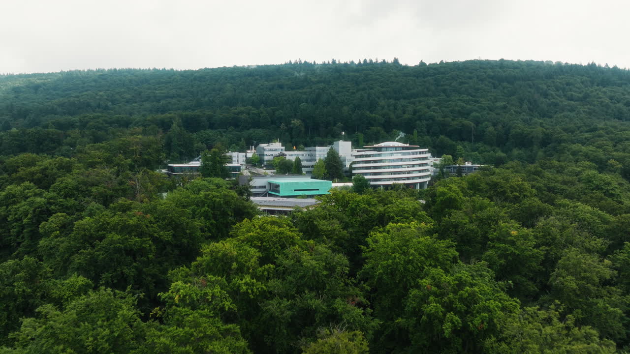 A drone flies above the treetops and moves forward toward the DKFZ complex, revealing the round tower, the green-roofed buildings, and the forested hillside