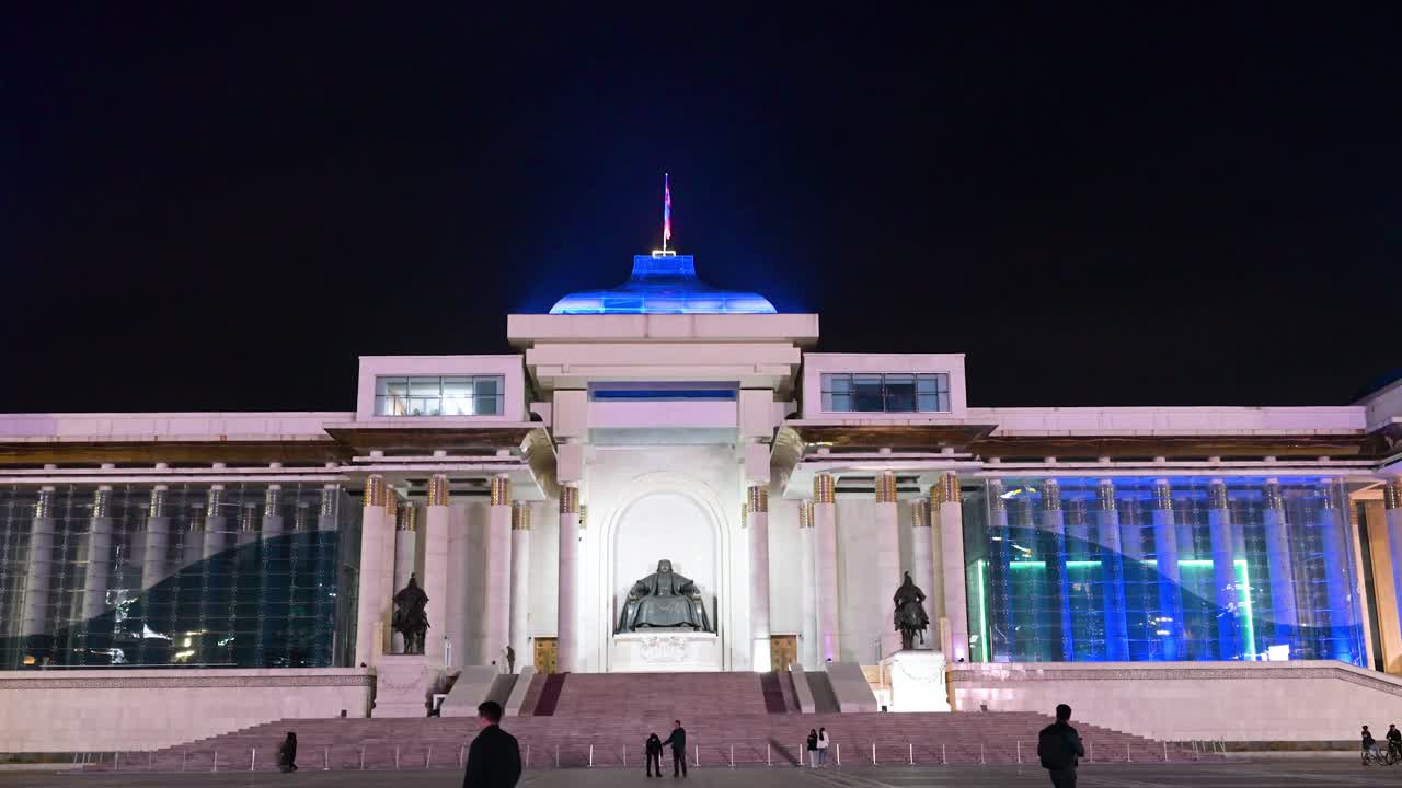In a peaceful nighttime scene, people stroll through Sukhbaatar Square, Ulaanbaatar, where the bronze statue of Genghis Khan stands before the Government Palace with a flag flying above.