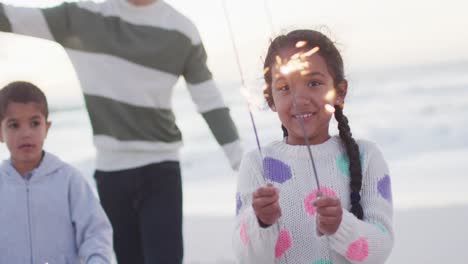 Portrait-of-happy-hispanic-girl-playing-with-sparklers-on-beach-with-family-at-sunset