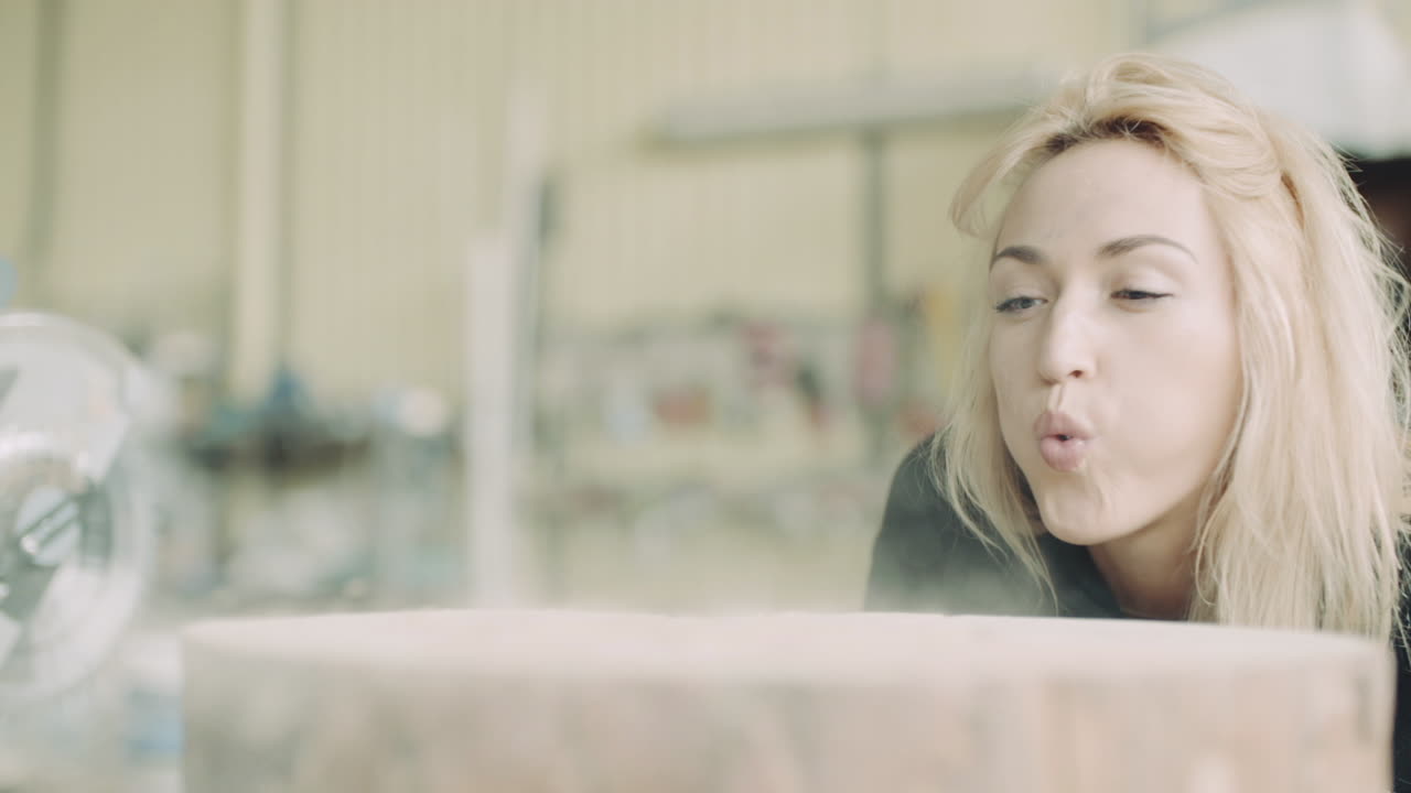 Young Woman Blowing Dust Off a Wooden Surface in a Workshop