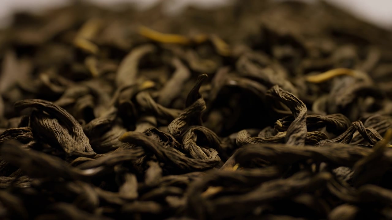 Close-up footage glides gently over loose dried tea leaves, bringing wrinkled curled pieces into focus while surrounding layers fade into soft blur under warm, subdued light.