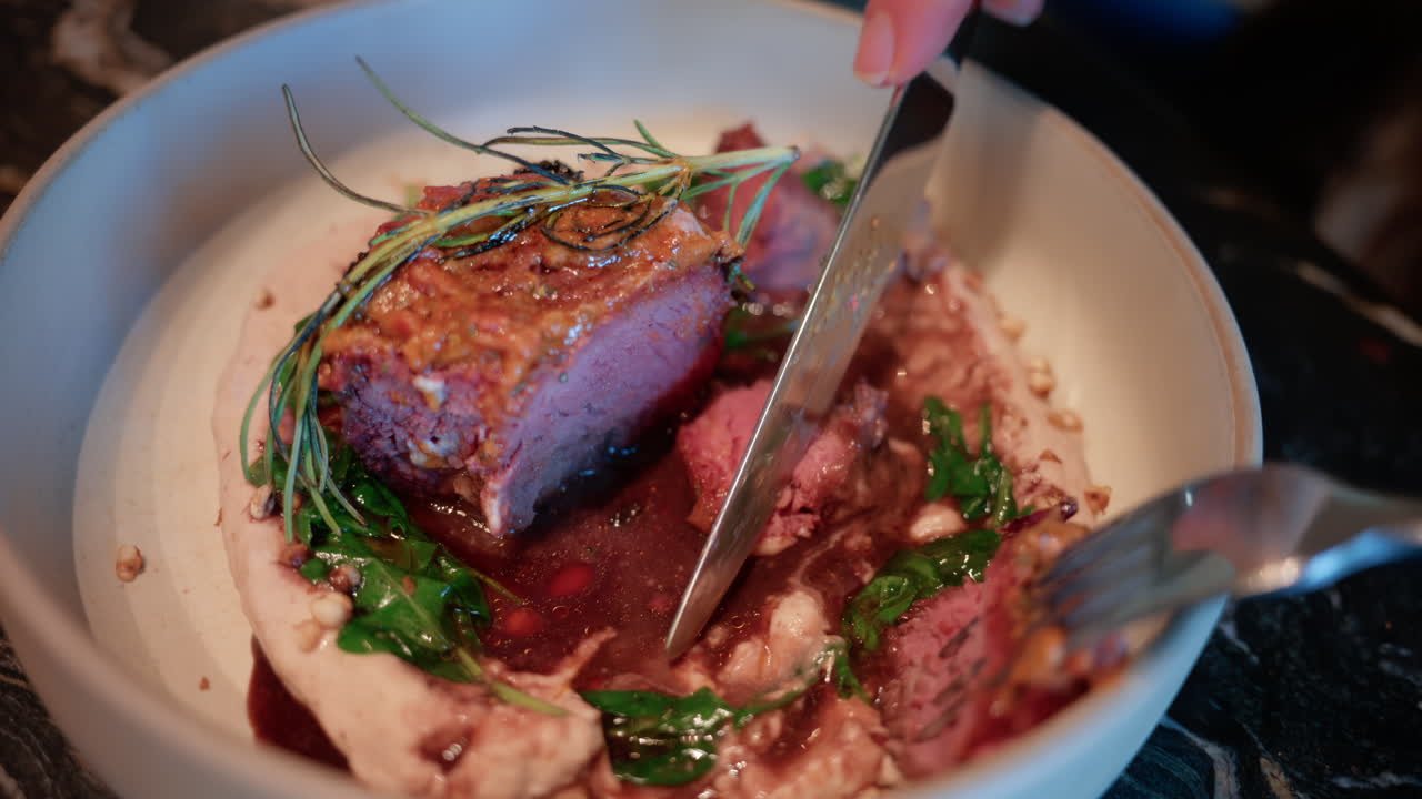 Close up of a woman's hand cutting into a gourmet meat dish served with sauce, greens, and rosemary garnish on a white plate