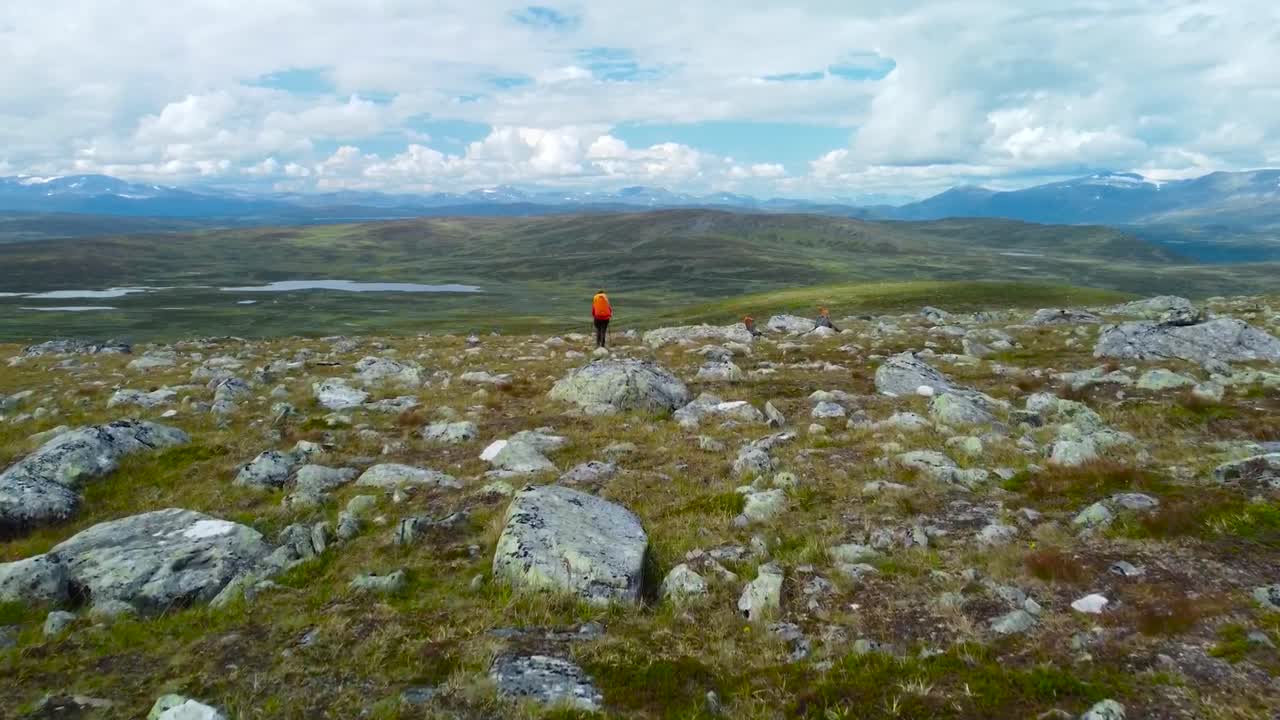 Aerial drone footage of two hikers or people walking and hiking on rocky and green grassy fields landscape in Sweden nature during cloudy day, hills and horizon visible in the background, white clouds