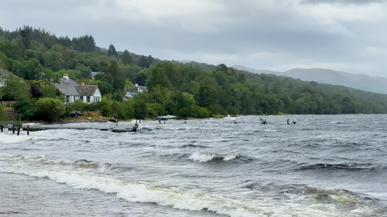 Choppy waves crash on Loch Ness shore, cloudy skies, green hills, static wide shot, daytime
